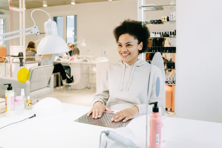 A client at a manicure station in a bright nail salon, curing lamps on the counter